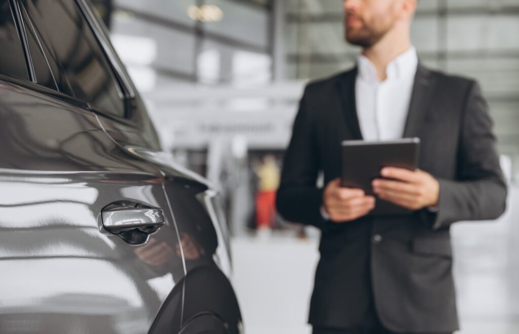Focus on the car and car seller in grey suit standing in defocus in car salon and holding tablet.